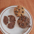 Two silicone chocolate cookie AirPods case covers with a real cookie on white plate, one caramel and one brown.