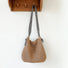 A straw bag with a long crossbody strap hangs on the wall, showcasing its details.