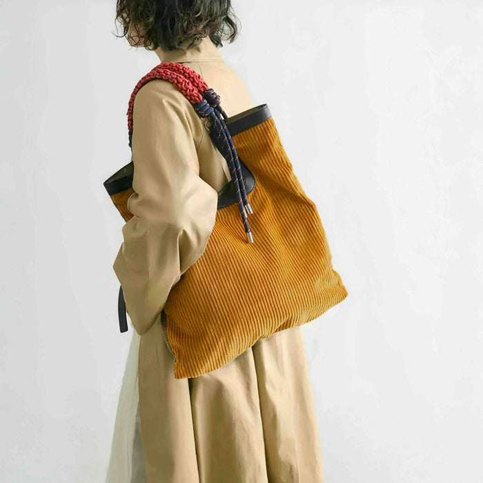 Side view of a woman wearing the designer gentle beam tote as a corduroy shoulder bag over a khaki trench coat.