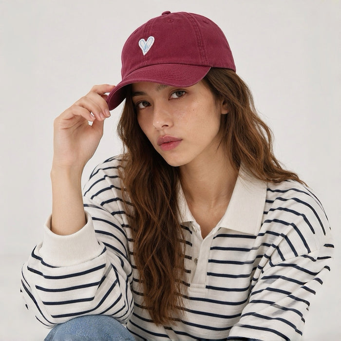 Female model wearing a burgundy cotton baseball cap with white heart embroidery paired with a striped polo.