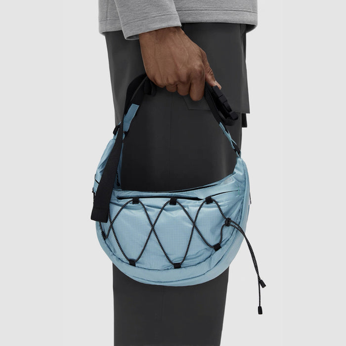 A person carrying a light blue airy nylon crescent sling bag by its adjustable strap, showcasing the bungee cord lattice.