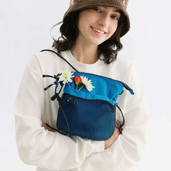 Smiling woman with a blue nylon Mesh Vogue Crossbody Bag, featuring a zip-top and front mesh pocket with flowers.