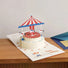 Front view of Pop-up carousel greetings card with red and white striped canopy, white horses, and Happy Birthday text.
